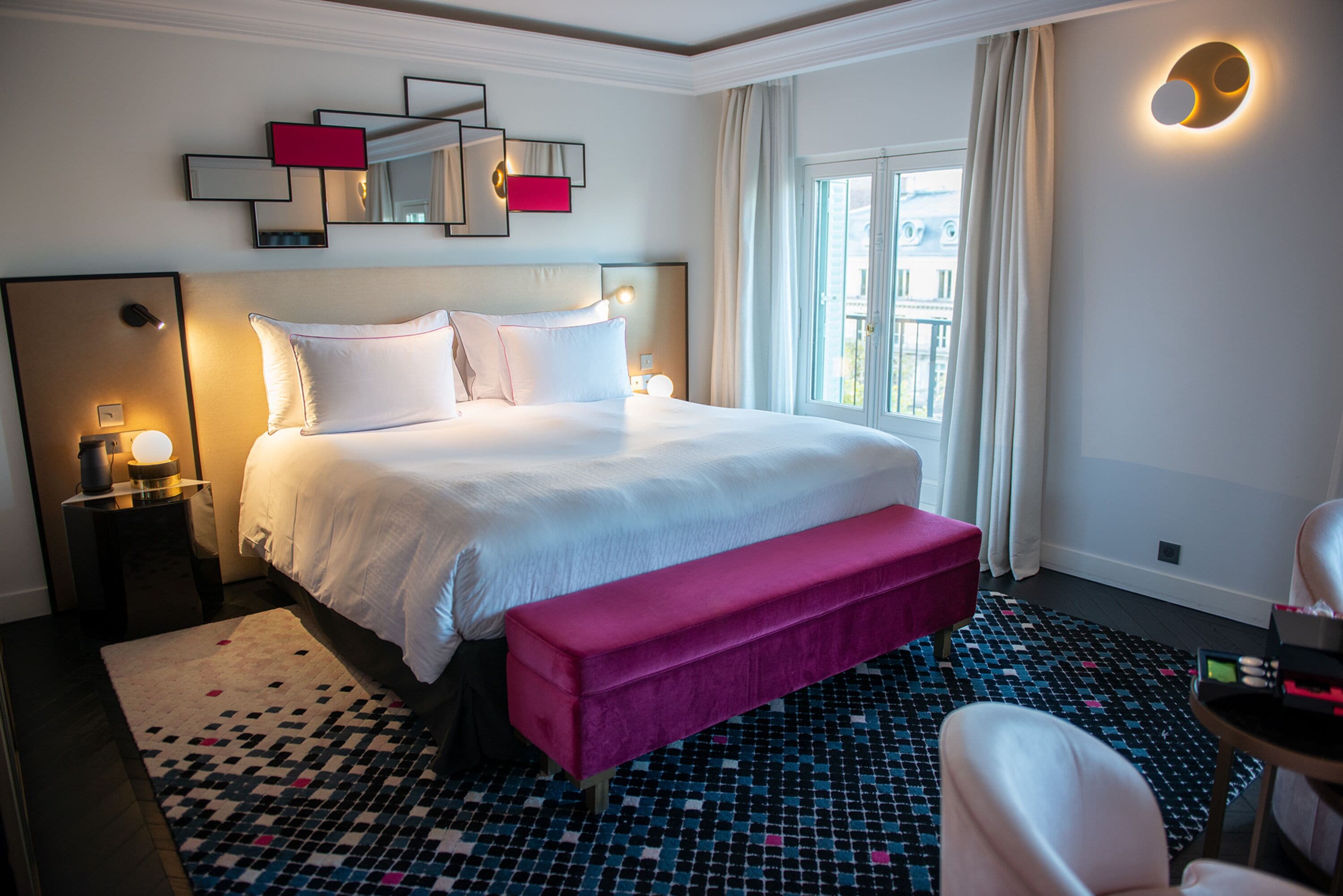 Ein Hotelzimmer mit einem großen Bett, einer rosafarbenen Bank, einem gemusterten Teppich und einem Nachttisch mit Lampe.