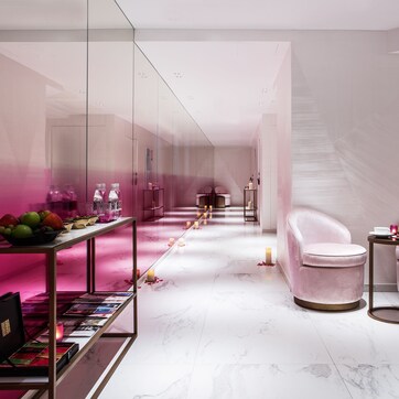 A modern interior with a marble floor, pink upholstered chairs, a glass display table with magazines, and a pink-lit corridor.