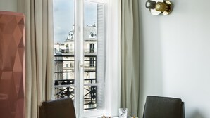 Quarto com vista para edifícios através de uma grande janela, duas cadeiras, uma mesa pequena e um abajur singular.