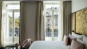 Habitación de hotel con una cama grande, una mesita con un florero y vistas a edificios a través de las ventanas.
