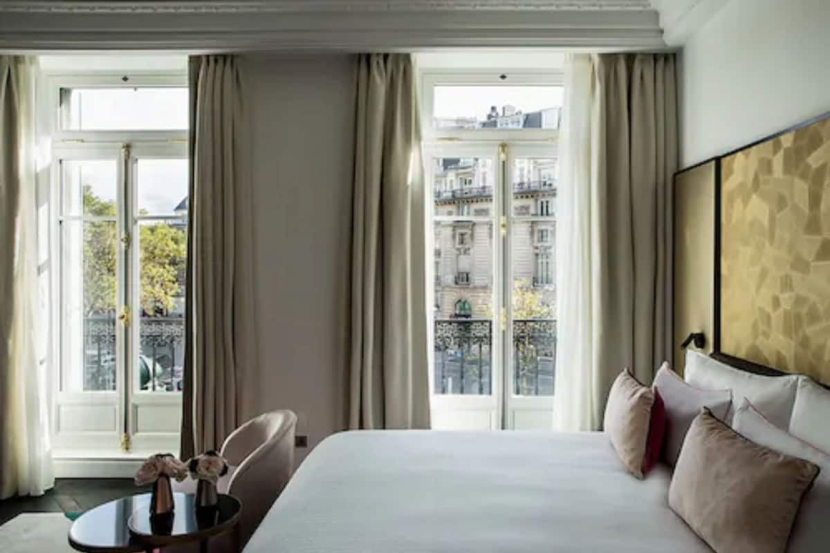 A hotel room with a large bed, a small table with a vase, and a view of buildings through the windows.