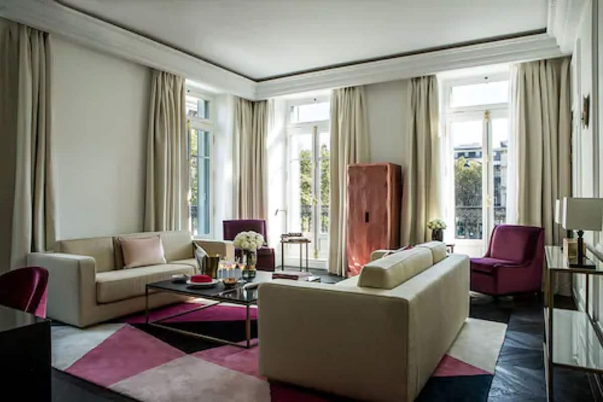 A modern living room with a beige sofa, a coffee table, and a red chair. There is a large window with curtains, and a view of the city.