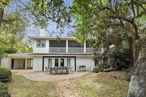 A two-story house with a covered patio, a dining area with chairs and a table, and a large tree in the foreground.