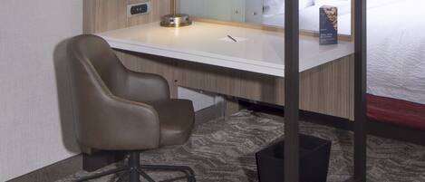 A hotel room with a desk, chair, bed, and a small trash bin.