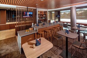 A modern bar area with a wooden ceiling, a television screen, and a bar stocked with various bottles. There are several wooden tables and chairs arranged for dining. - SpringHill Suites by Marriott Newark Downtown (Newark)