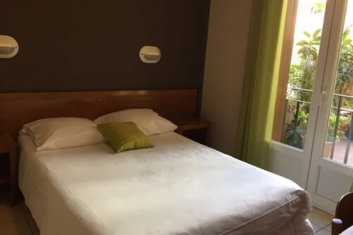 A hotel room with a bed, a nightstand, a window with curtains, and a view of greenery outside.