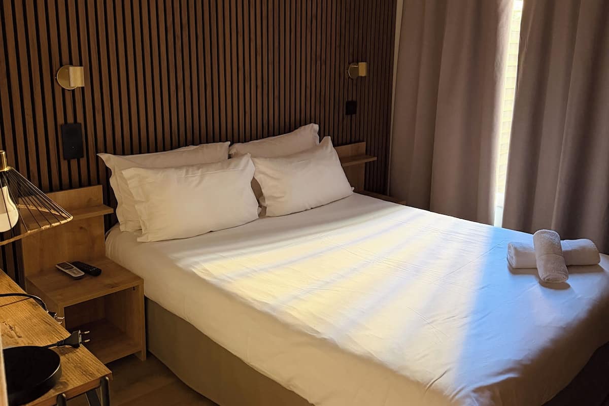 A hotel room with a large bed, wooden panel wall, bedside lamps, a nightstand, and a window with curtains.