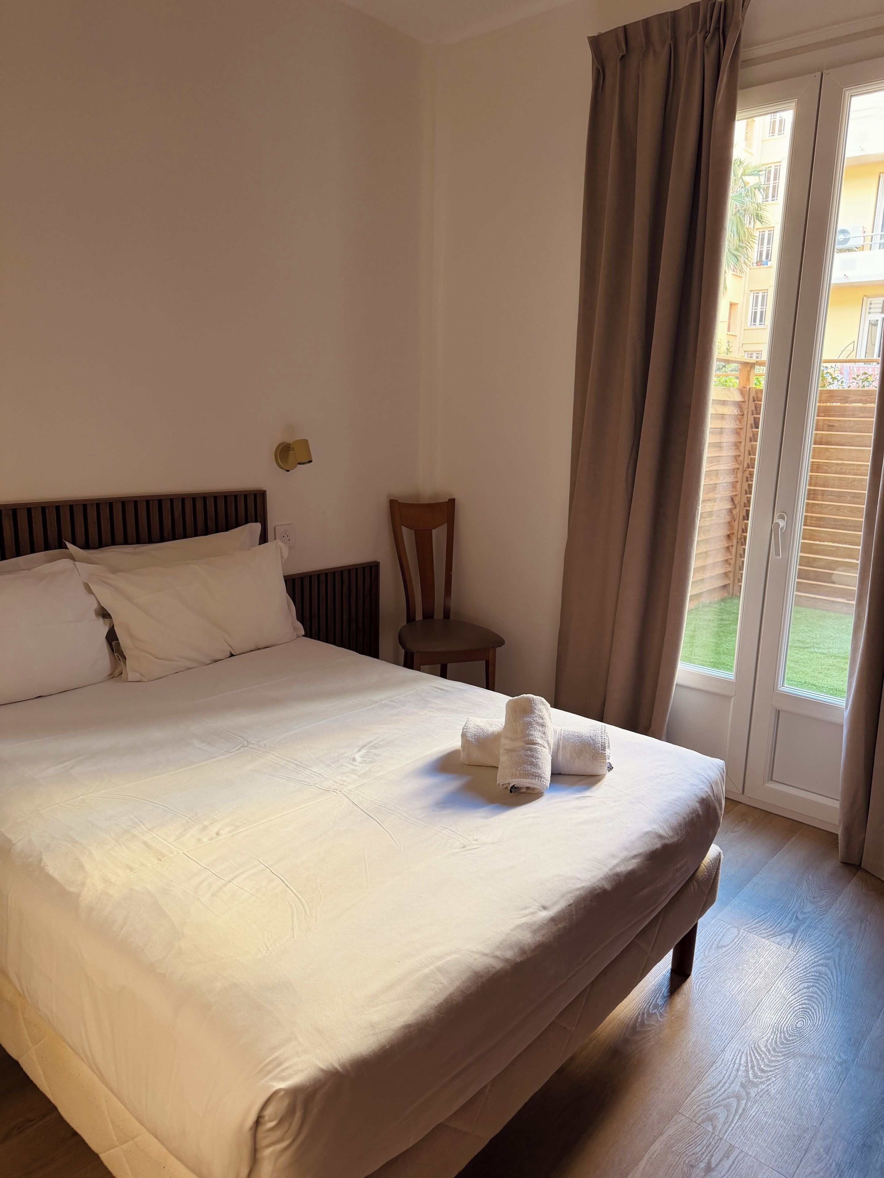 Ein ordentlich gemachtes Bett mit einem gefalteten Handtuch, ein Holzstuhl und ein Fenster mit Vorhängen.