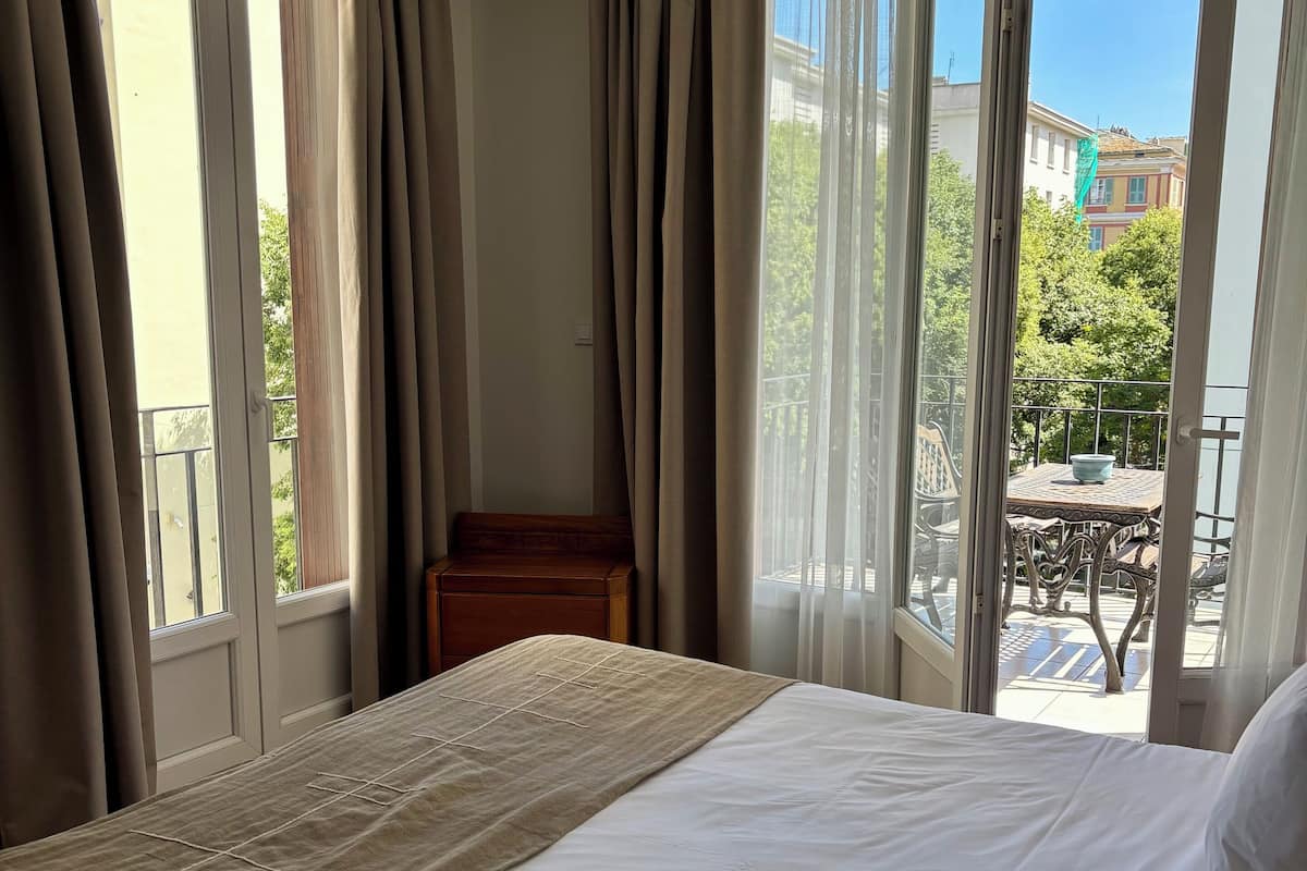 A hotel room with a large bed, a wooden nightstand, and a view of the outdoor balcony and buildings.