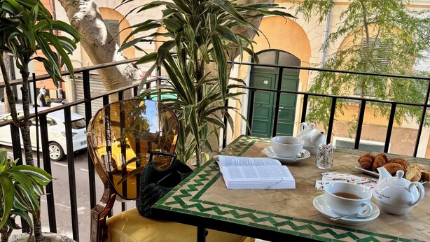 A table with a green patterned tablecloth, a chair, a potted plant, a book, a cup of tea, a teapot, and a plate with croissants. - Hôtel Bonaparte (Bastia)