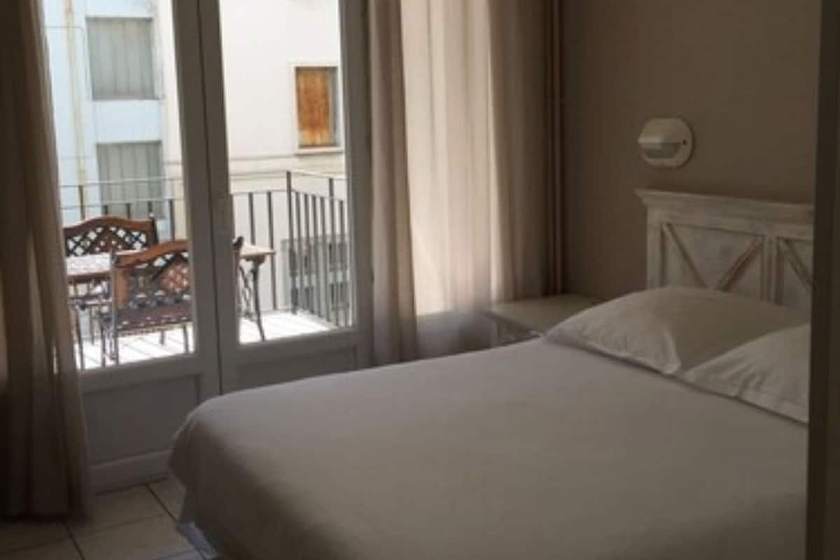 A hotel room with a bed, a window with curtains, a view of the outside balcony, and a wall-mounted light fixture.