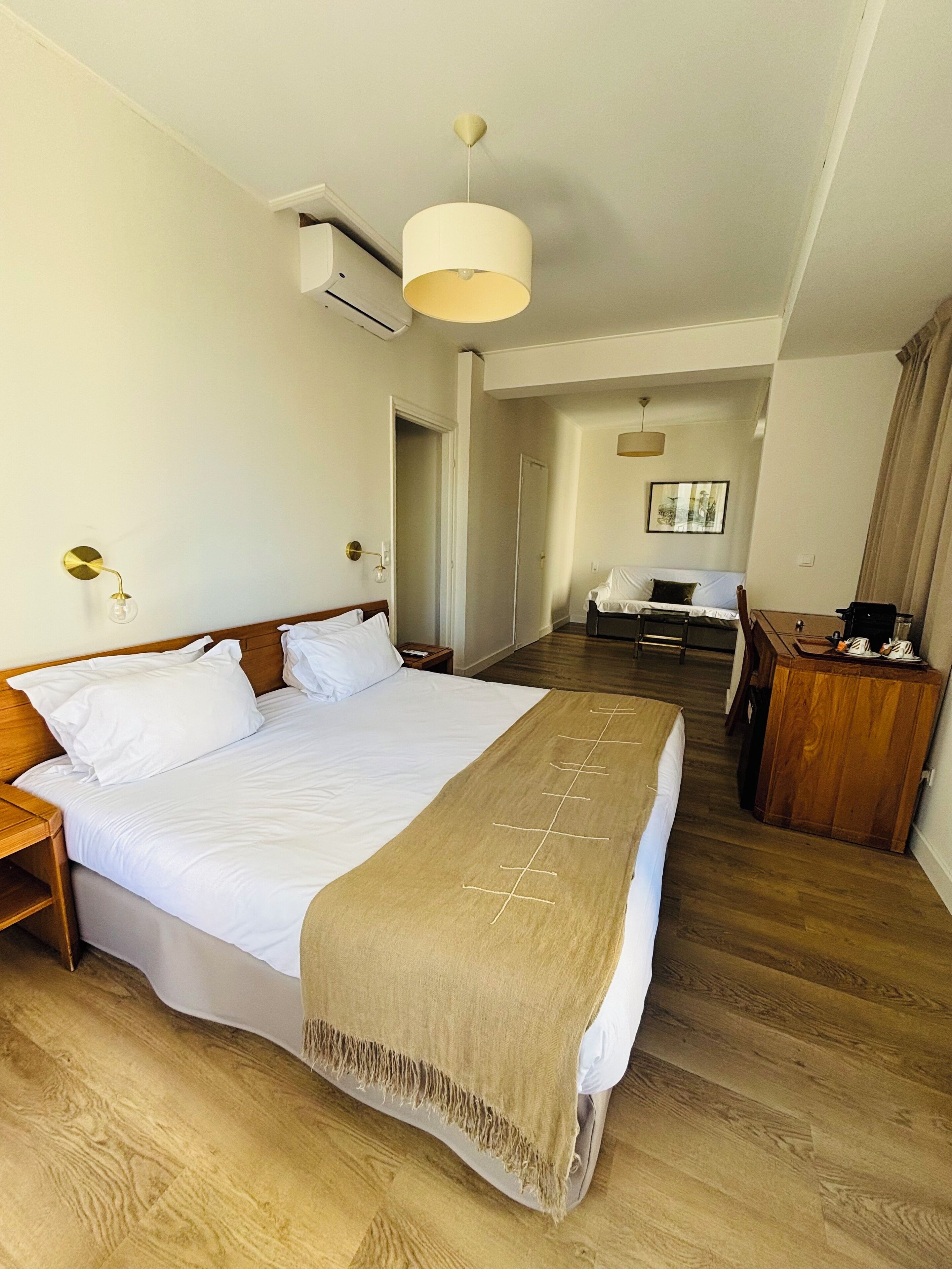 A hotel room with a large bed, a wooden nightstand, a desk, and a chair. There is a door leading to another room and a window with curtains.