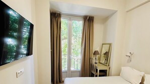 A hotel room with a bed, a desk with a chair, a TV mounted on the wall, and a window with curtains. - Hôtel Bonaparte (Bastia)