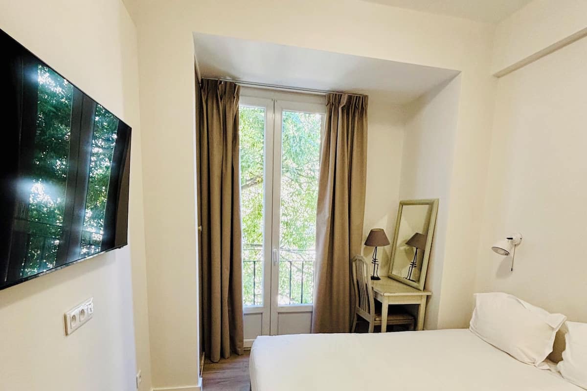 A hotel room with a bed, a desk with a chair, a TV mounted on the wall, and a window with curtains.