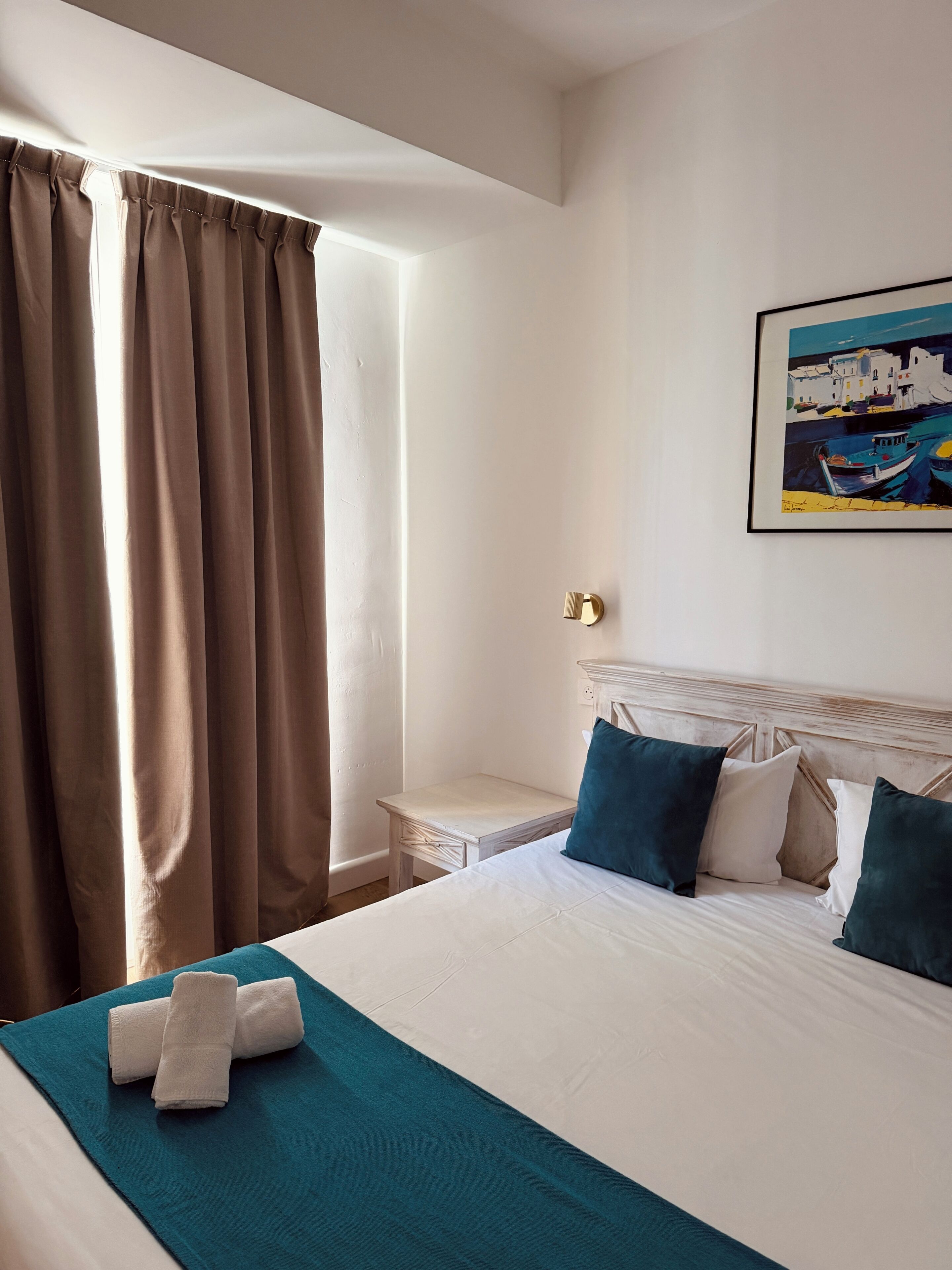 A hotel room with a bed, a nightstand, a painting, and a window with curtains.