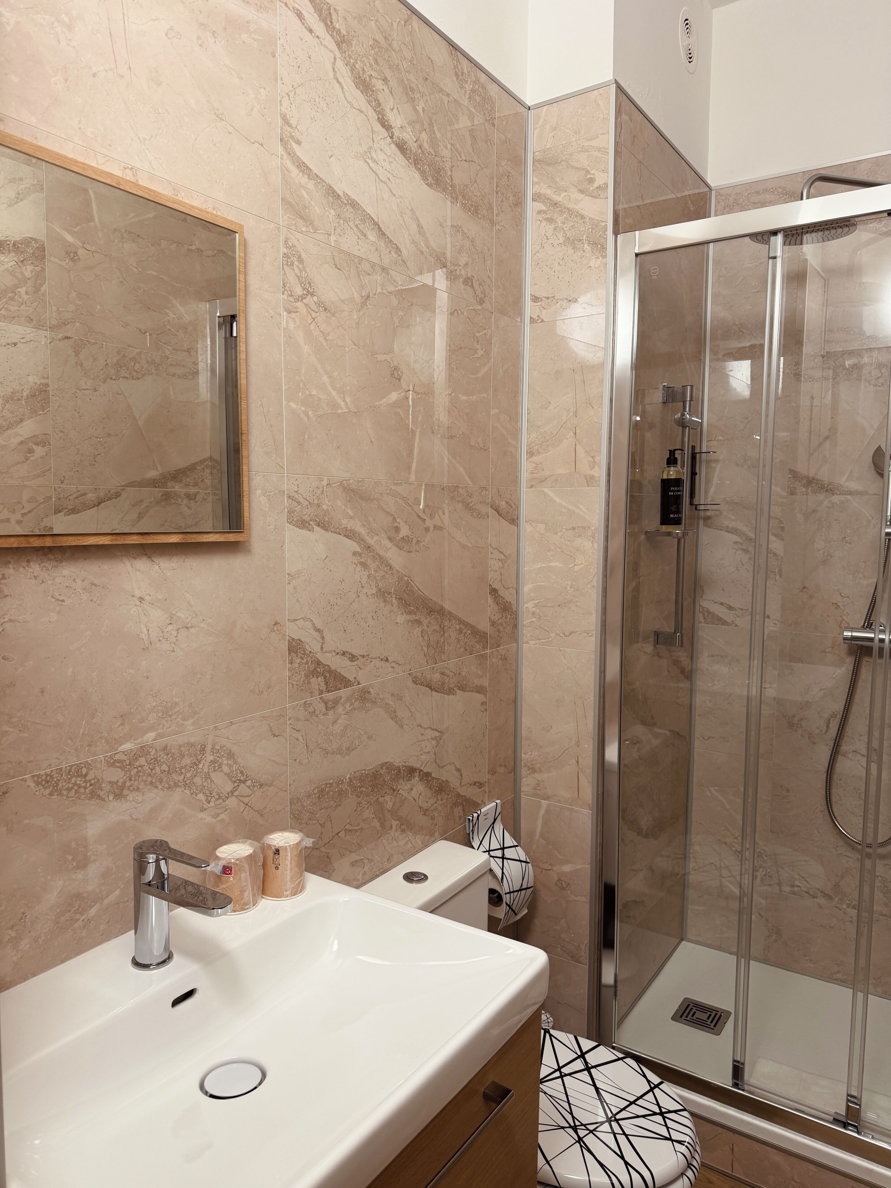 Ein Badezimmer mit Marmorwand, Waschbecken, Spiegel, Duschkabine aus Glas und Toilette mit geometrischem Design.