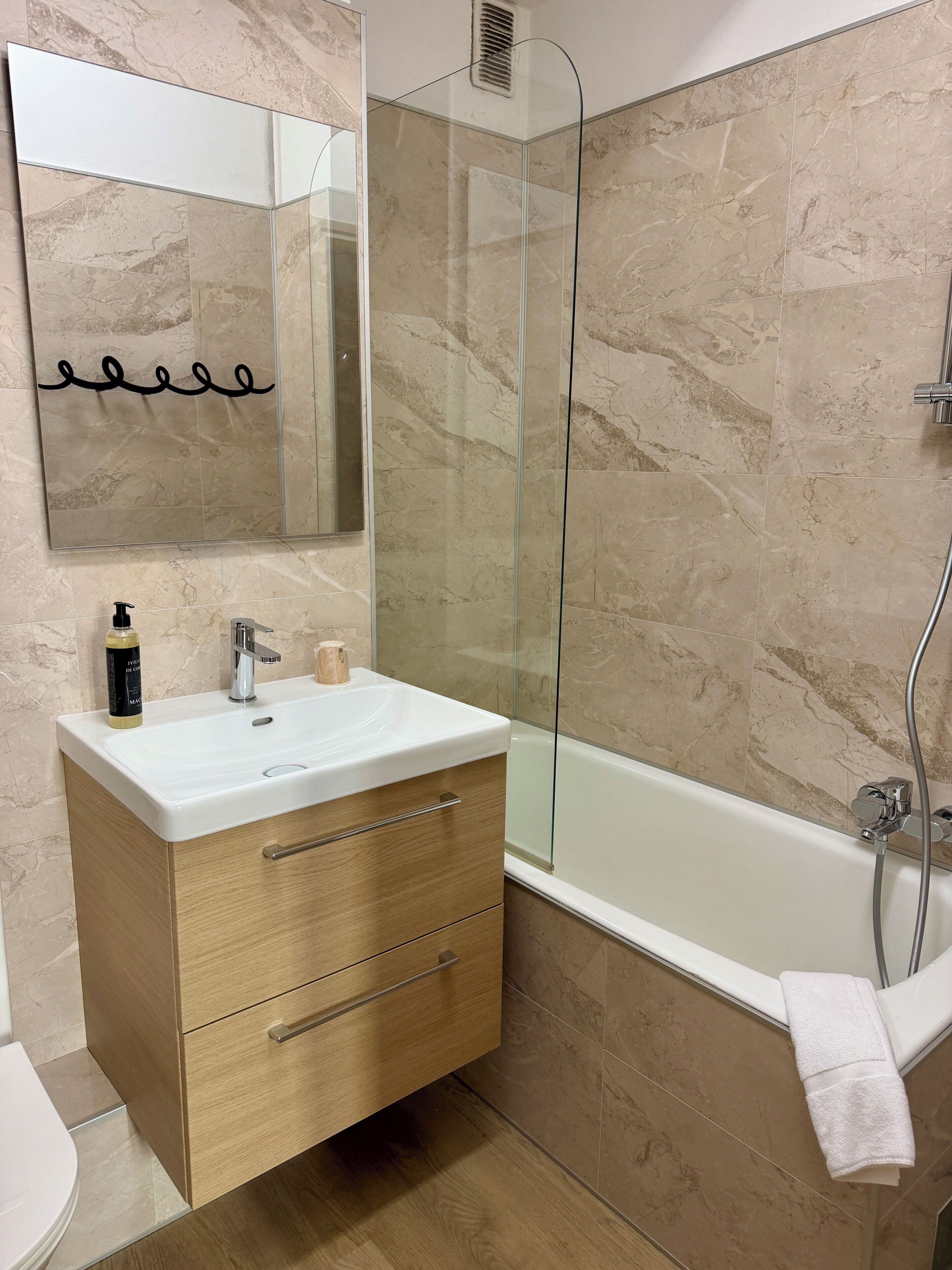 A bathroom with a marble-tiled wall, a glass-enclosed shower, a white sink with a wooden vanity, and a towel hanging on the wall.