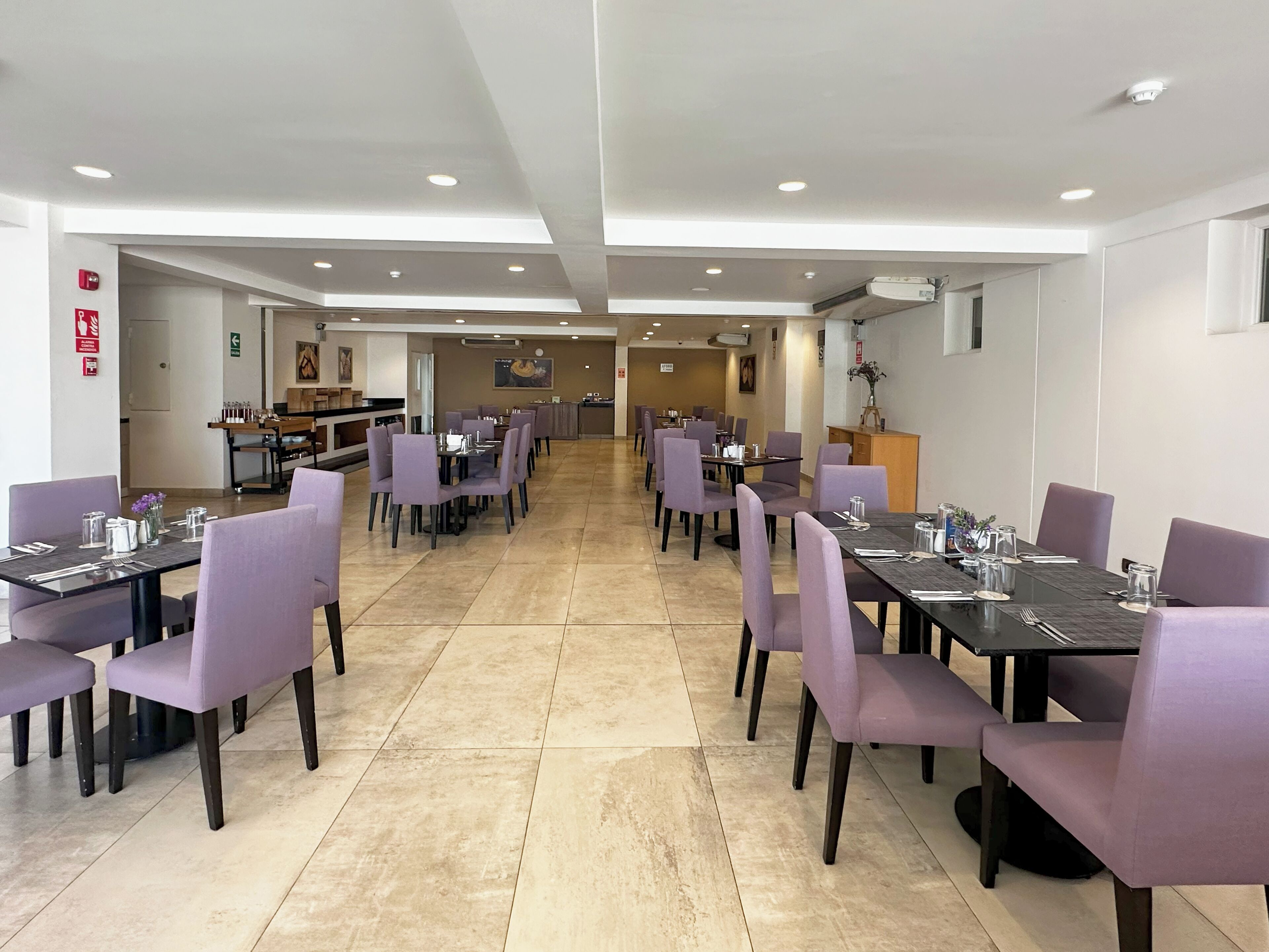 A hotel dining area with multiple tables and chairs, a kitchen in the background, and a fire extinguisher on the wall.