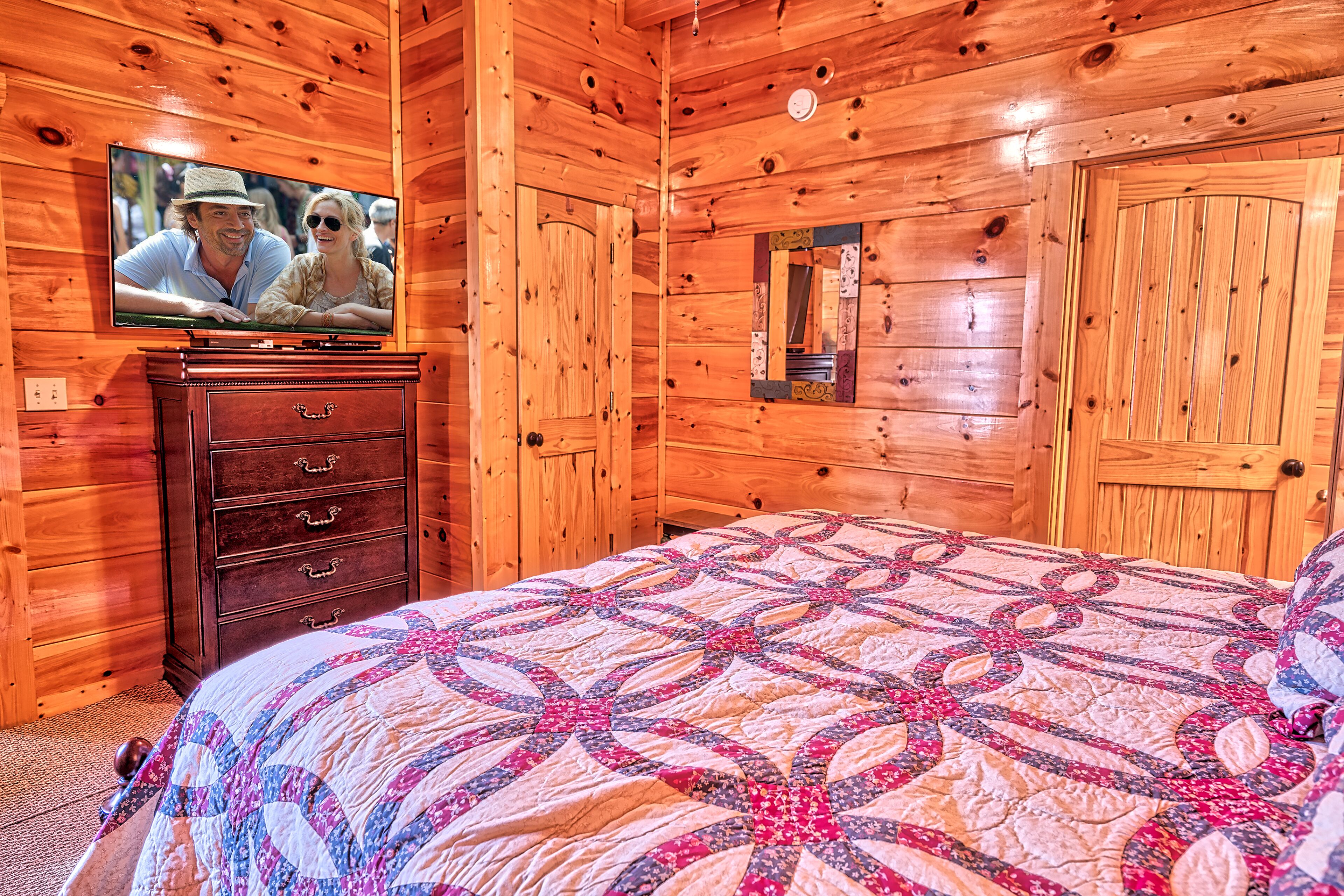 Ein Zimmer in einer Holzhütte mit einem Bett, einem Schminktisch, einem Fernseher und einem Spiegel.