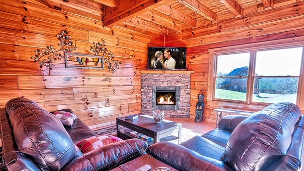 Interior acogedor de una cabaña de troncos con sofás de cuero, chimenea y un ventanal con vistas al exterior.