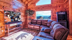 Interior de una cabaña de madera con un sofá de cuero, un televisor mostrando un partido de fútbol y vistas al exterior a través de una ventana.