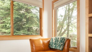 A room with a leather armchair and ottoman, a wooden shelf, and large windows with a forest view.