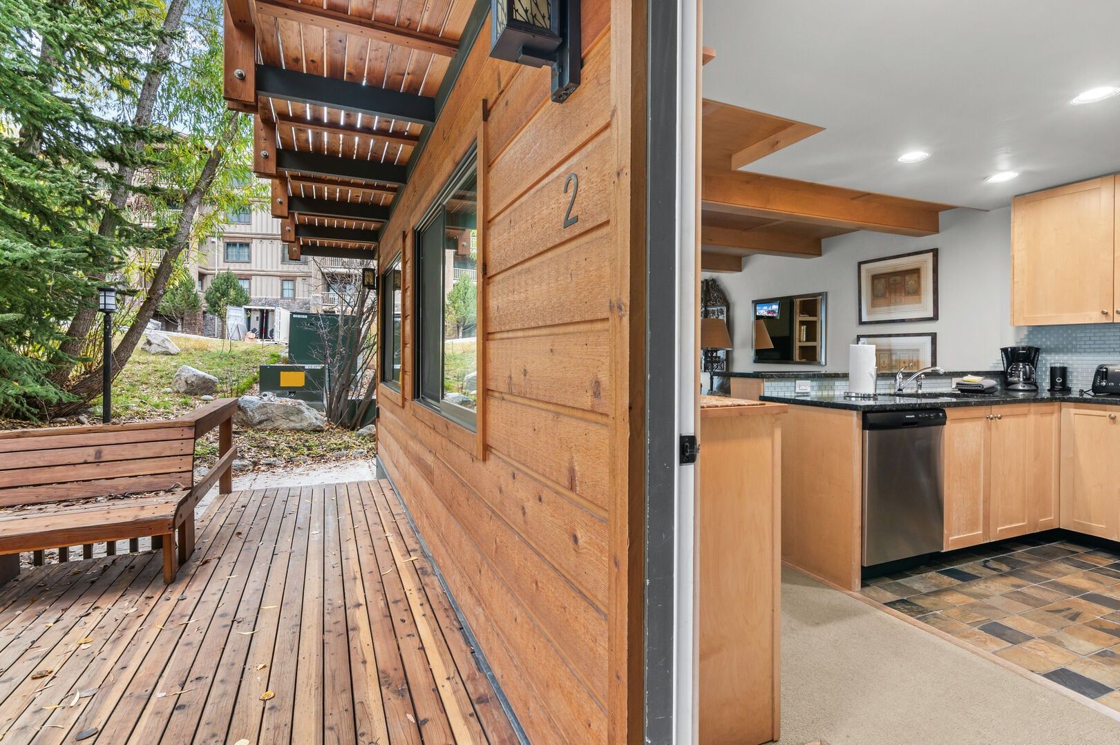 A wooden deck with a bench, a kitchen with wooden cabinets, and a view of a residential area.
