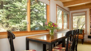 A dining area with a wooden table and chairs, a vase with flowers, and large windows with a forest view.