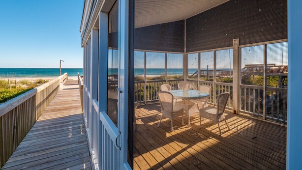 Une terrasse en bois offrant une vue sur l’océan, un espace extérieur couvert avec des chaises et une table, et une grande fenêtre donnant sur la plage.