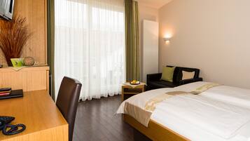 A hotel room with a wooden desk, a bed with white linens, a small side table with a fruit bowl, and a green curtain.