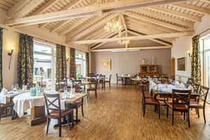 Uma sala de jantar ampla com vigas de madeira e um candelabro, com várias mesas e cadeiras, e grandes janelas com cortinas.