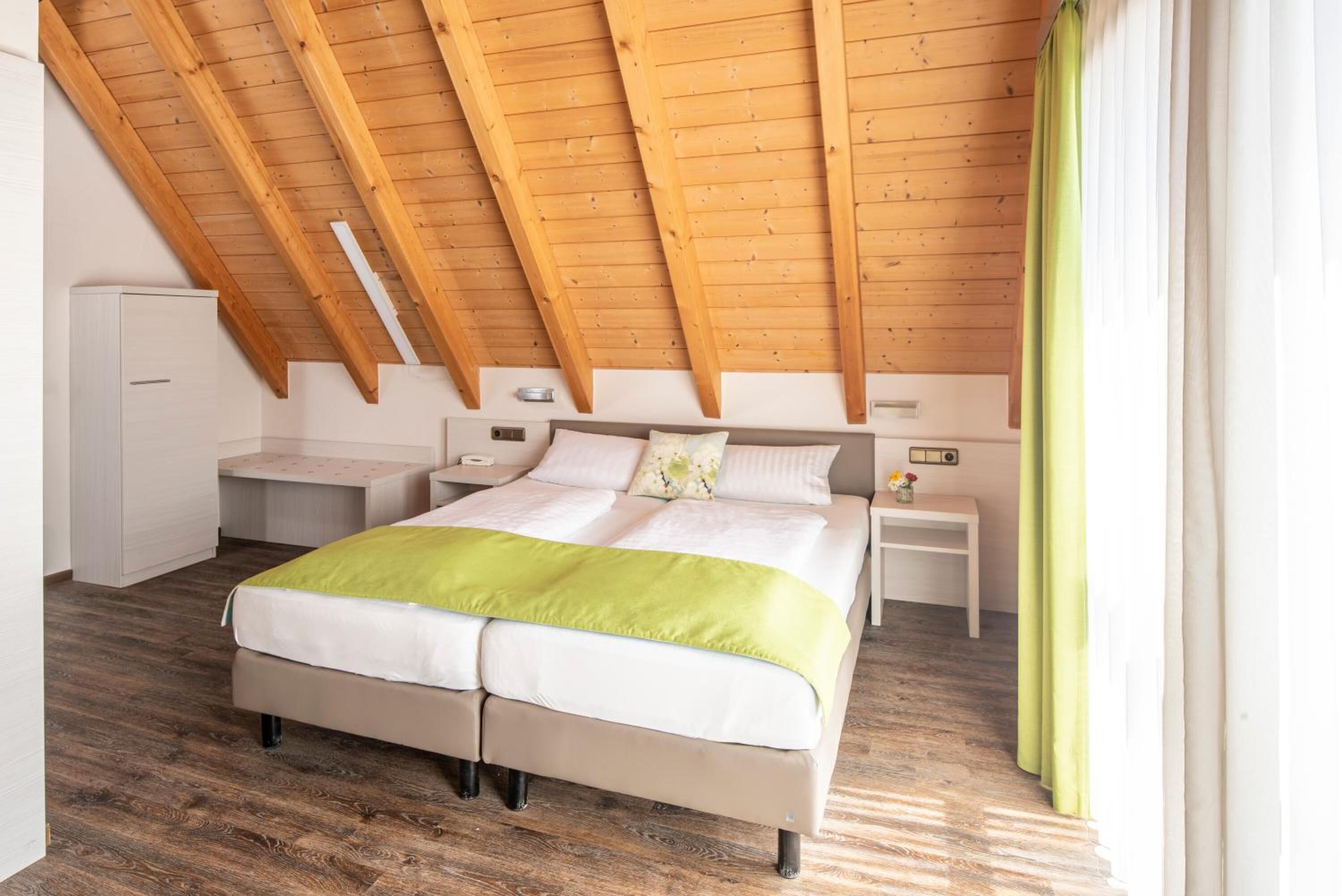Een slaapkamer met een schuin houten plafond, een bed met een groene deken, nachtkastjes en een raam met gordijnen.