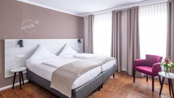 Quarto de hotel moderno com uma cama grande, uma mesa redonda pequena, uma cadeira e uma janela com cortinas.