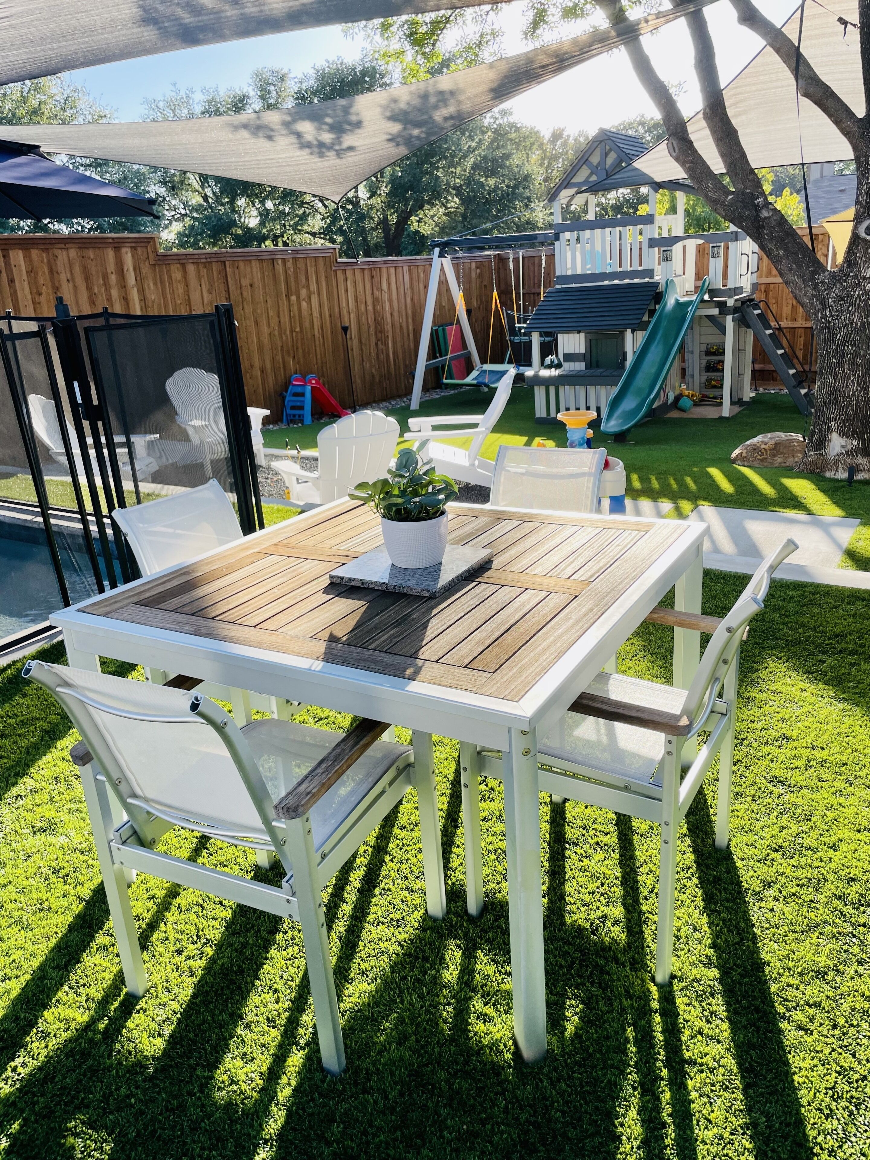 Een achtertuin met een houten tafel en witte stoelen, een kleine tuin en een glijbaan.