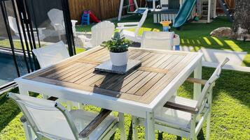 Een achtertuin met een houten tafel en witte stoelen, een kleine tuin en een glijbaan.