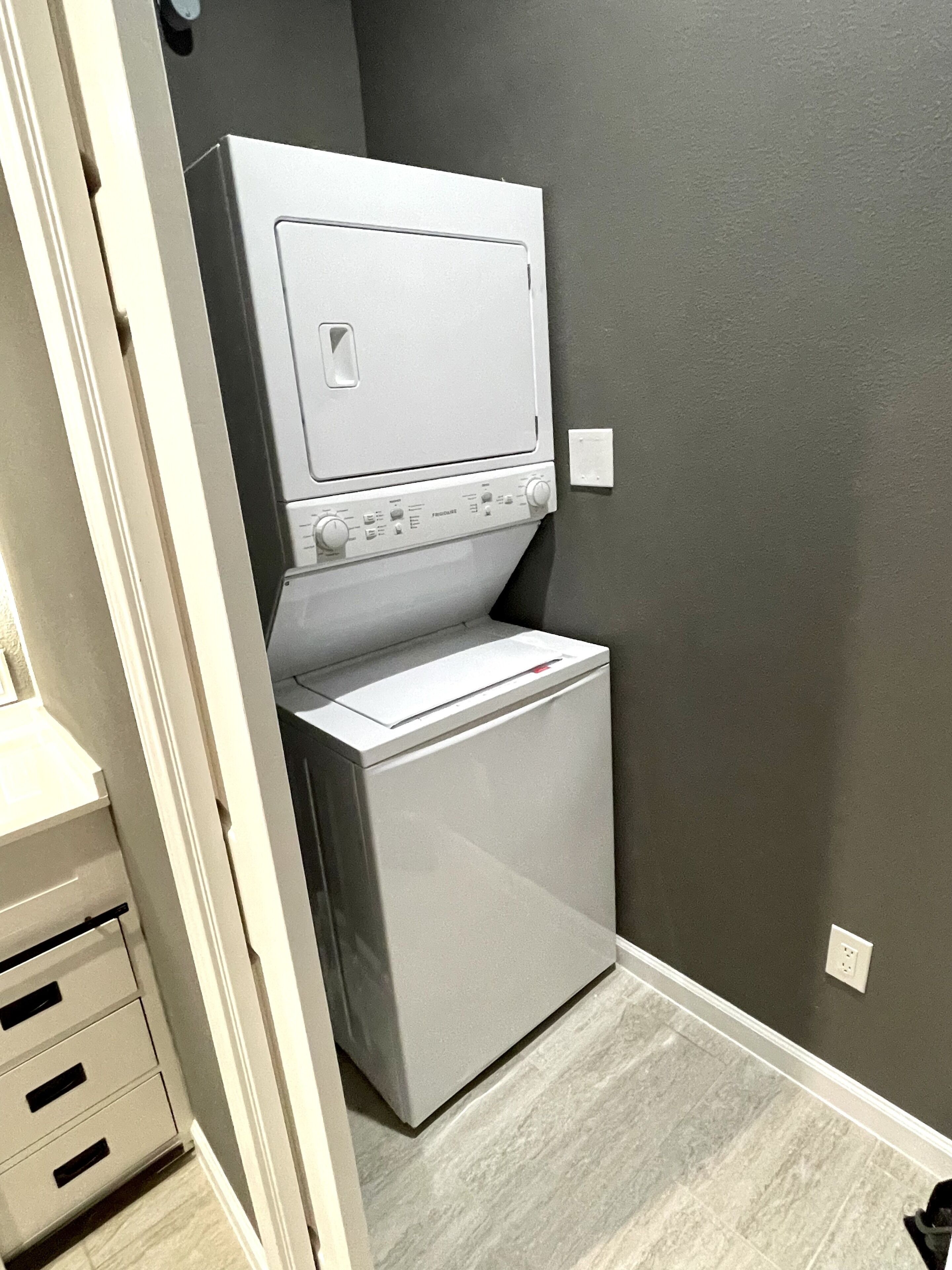 A compact laundry room with a front-loading washer and dryer, a white cabinet with drawers, and a gray wall.