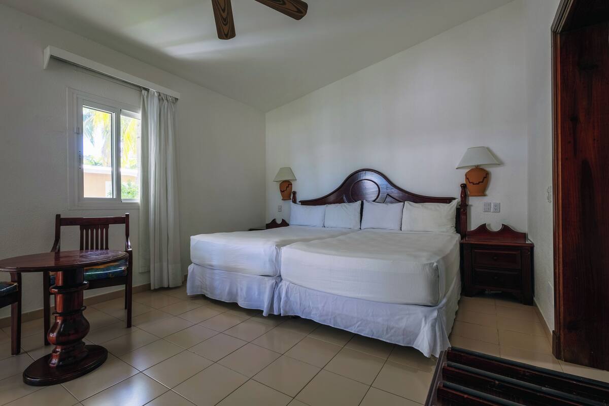 A double bed with white bedding, a wooden nightstand with a lamp, a wooden chair, and a window with curtains.