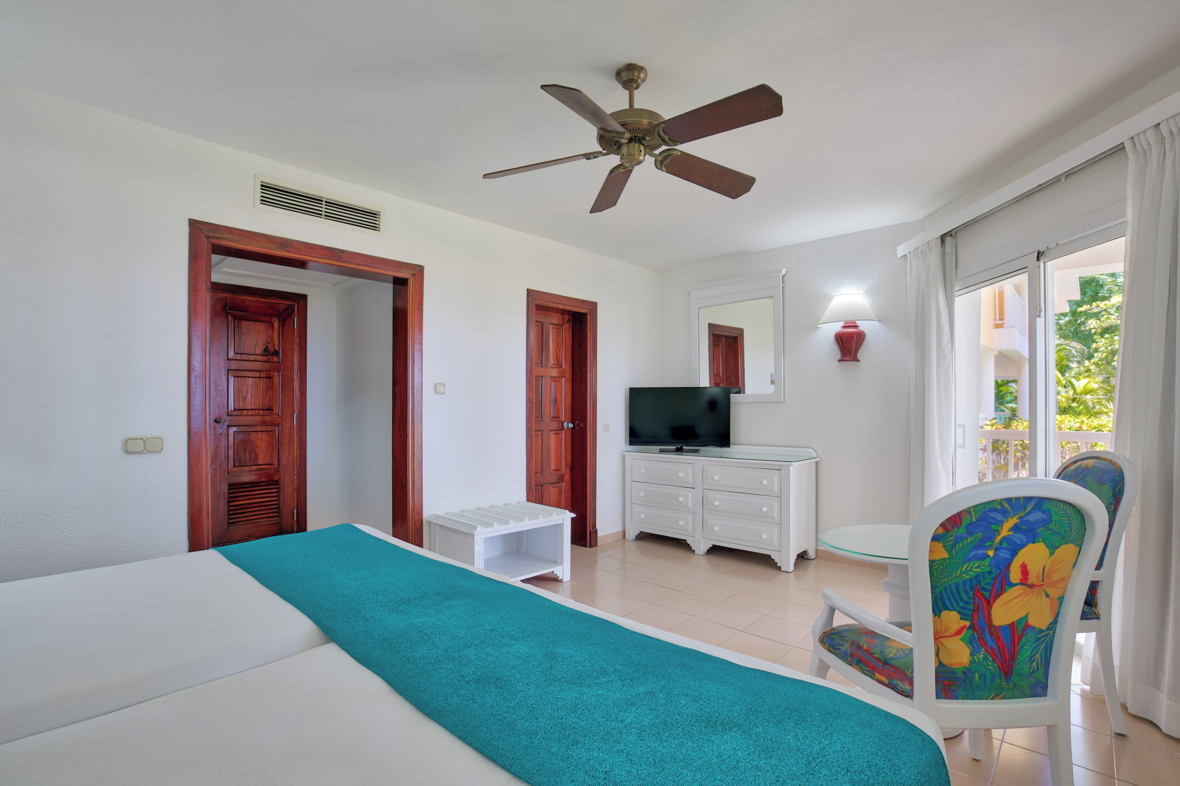 A bedroom with a bed, a TV, a ceiling fan, a small table, and a chair.