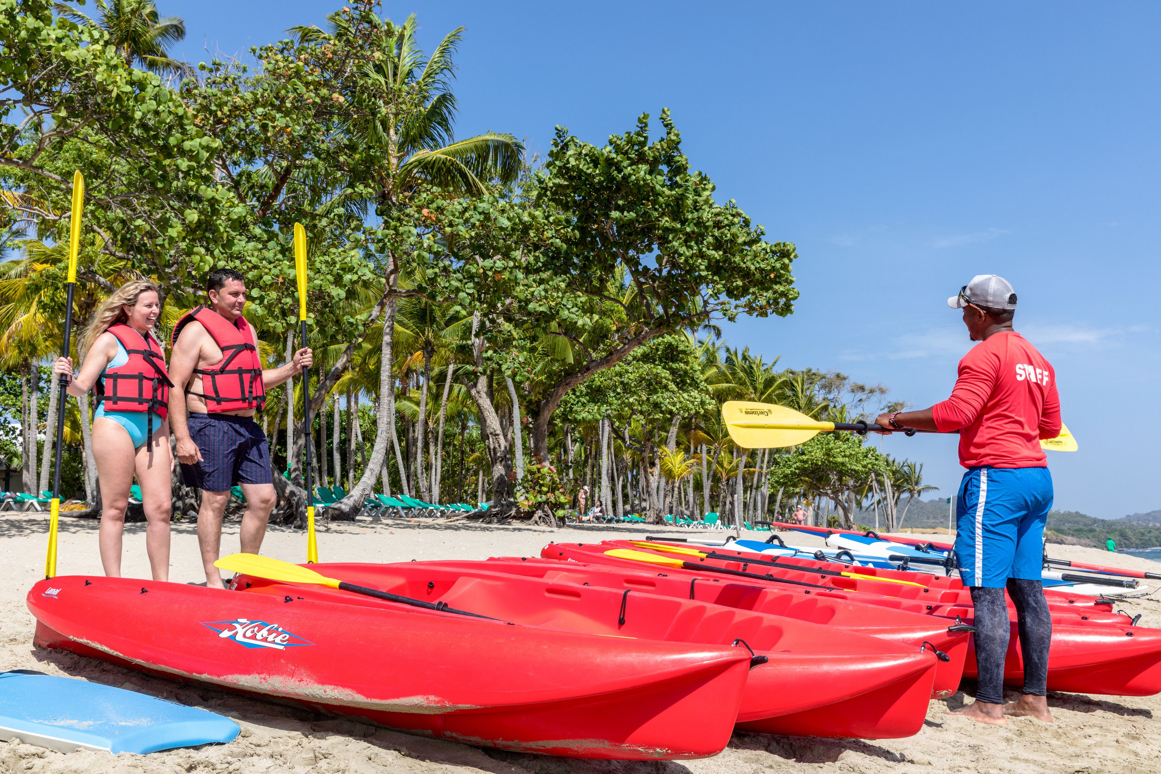 Una playa con kayaks rojos alineados y una persona con camisa roja de pie, sosteniendo un remo.