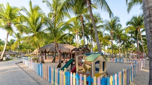 A beachside playground with colorful play structures, surrounded by palm trees and a thatched-roof hut.
