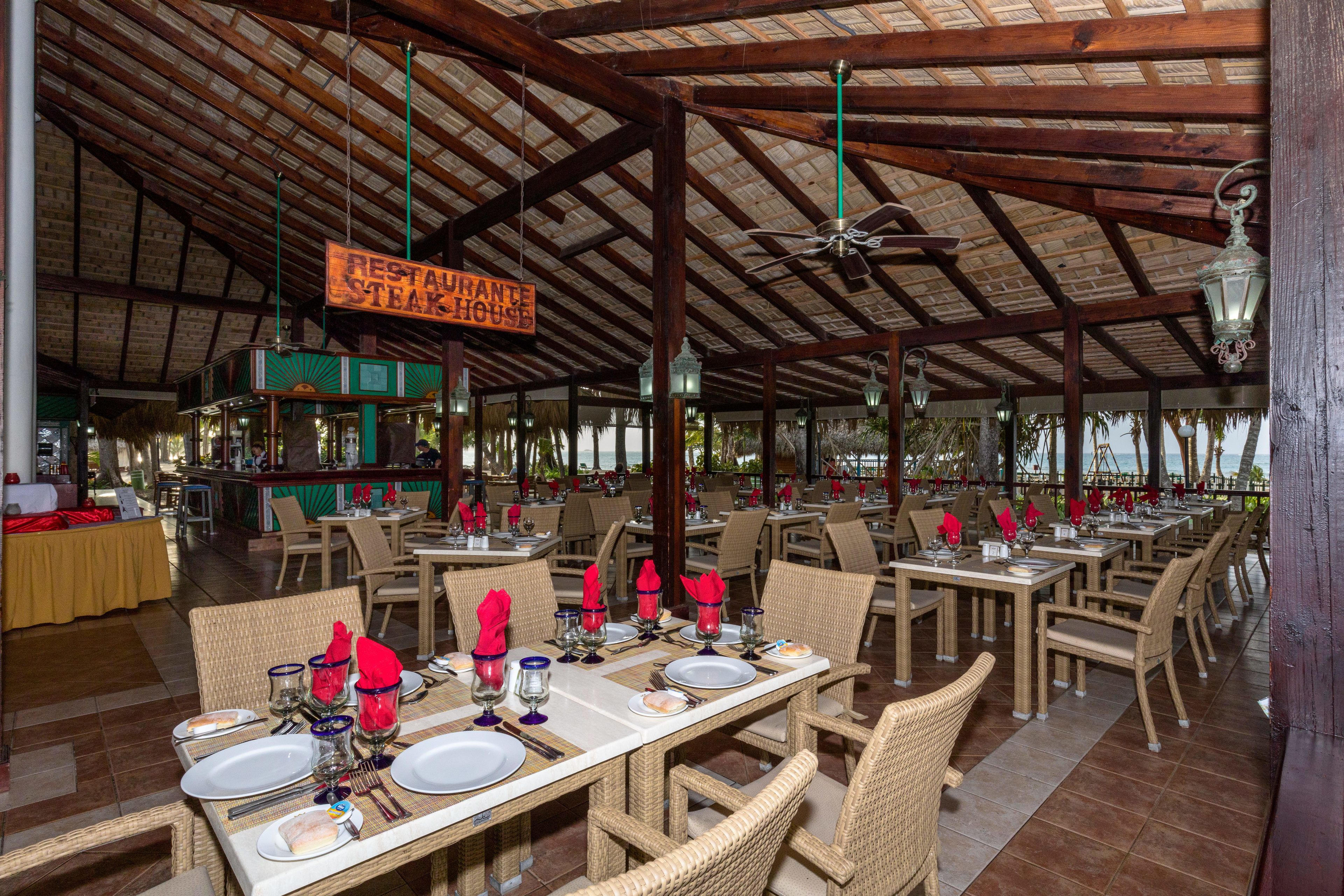 A restaurant with wooden beams and a sign for steakhouse. Tables are set with plates, glasses, and red napkins.