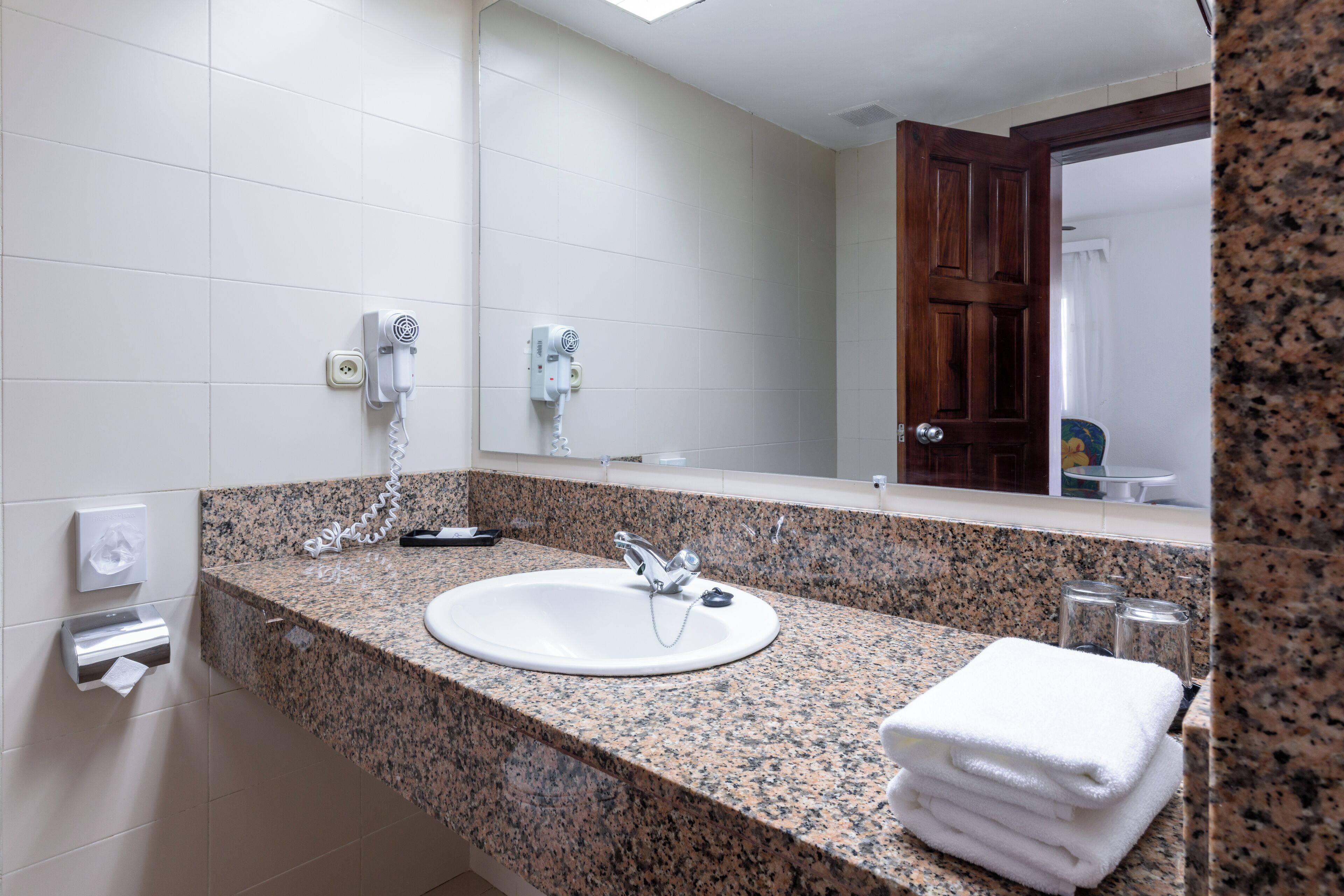 Bagno con top in granito, lavandino bianco, specchio, asciugacapelli e porta asciugamani.