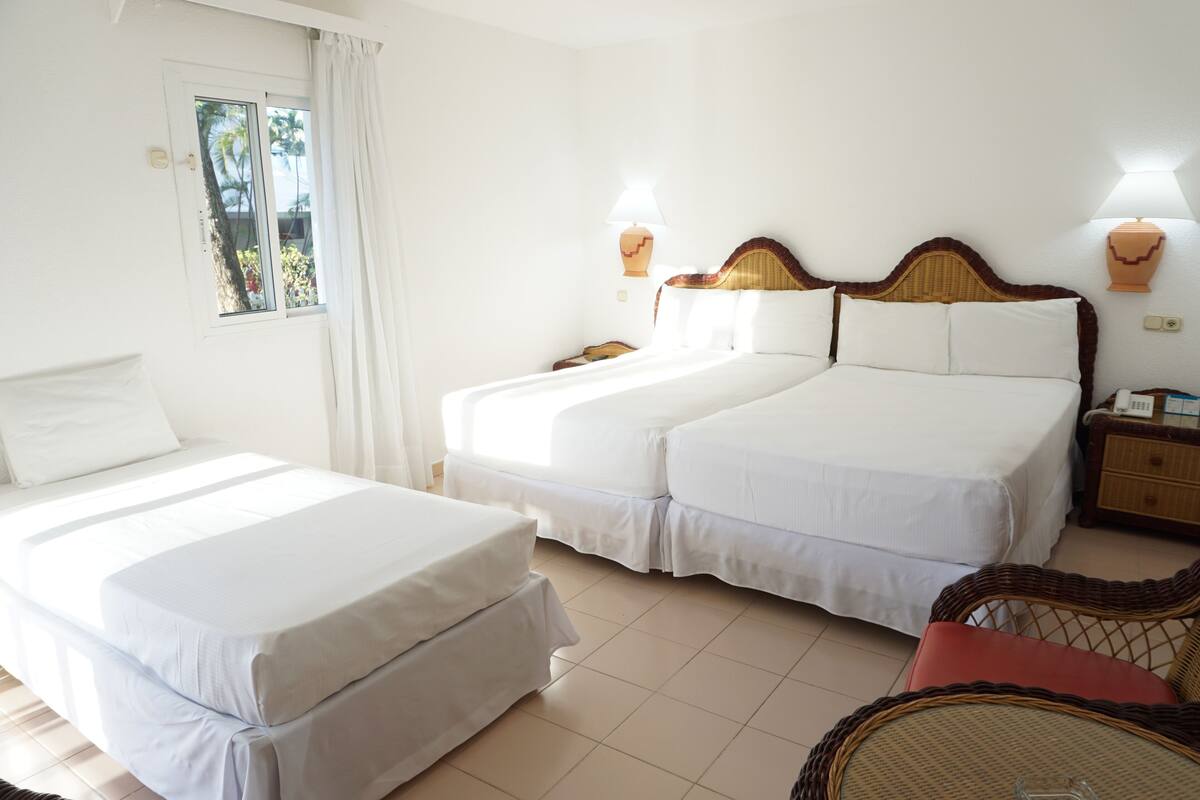 A hotel room with two beds, a window with curtains, a wicker chair, and two wall-mounted lamps.