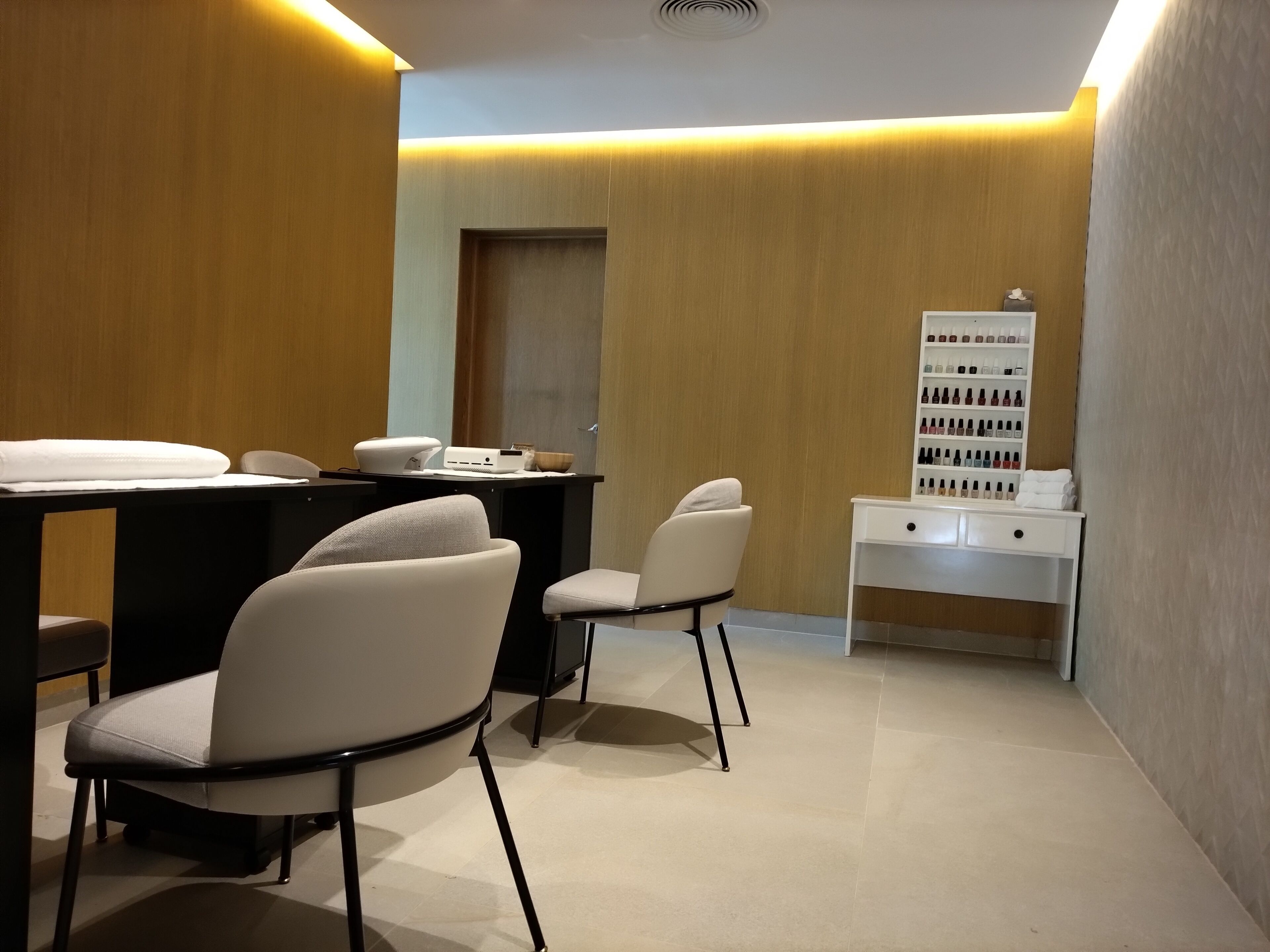 A modern, well-lit room with a reception desk, two chairs, a white console table with a display of nail polish, and a door in the background.