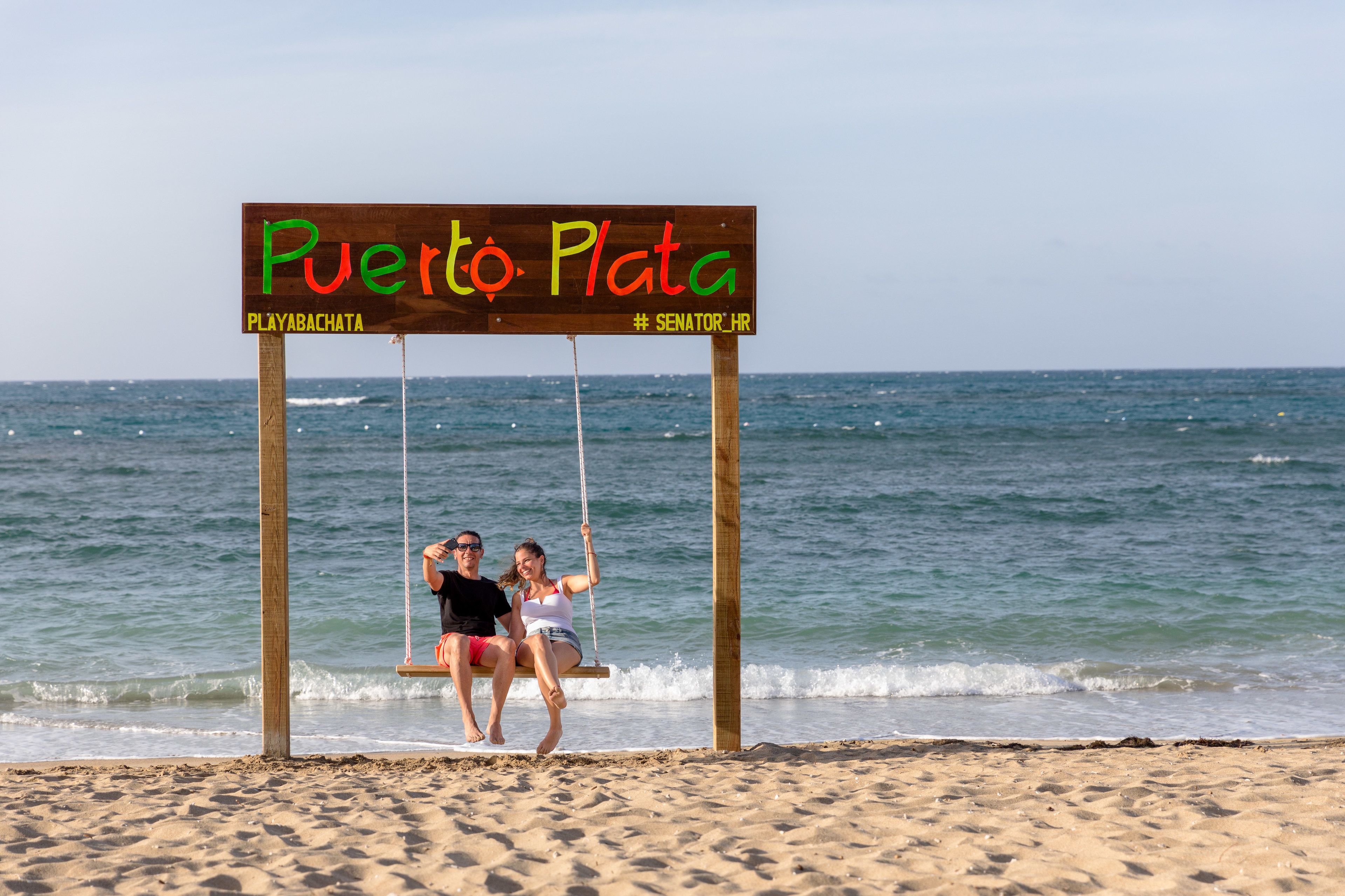 A wooden swing set with a sign that reads "Puerto Plata Playachata # Senator HR" on a beach with the ocean in the background.