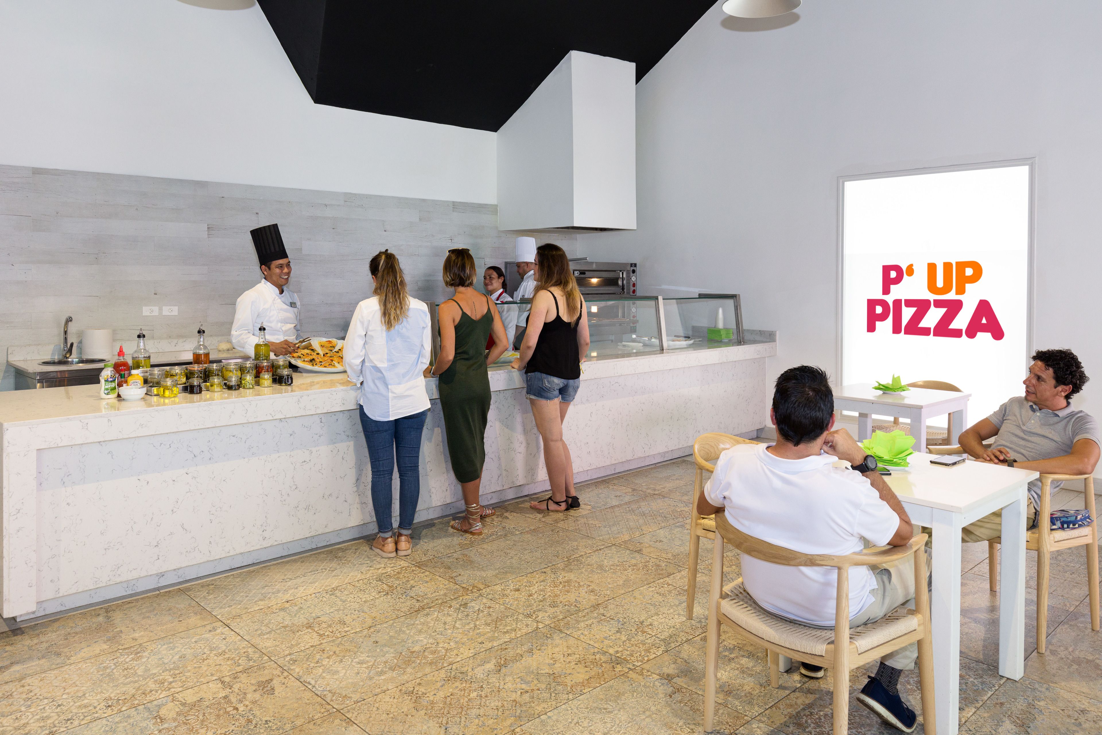 A modern kitchen area with a pizza counter, a large pizza sign, and a seating area with tables and chairs.
