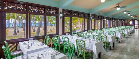 A restaurant with multiple tables and chairs, large windows with a view of palm trees and a beach, and ceiling fans.