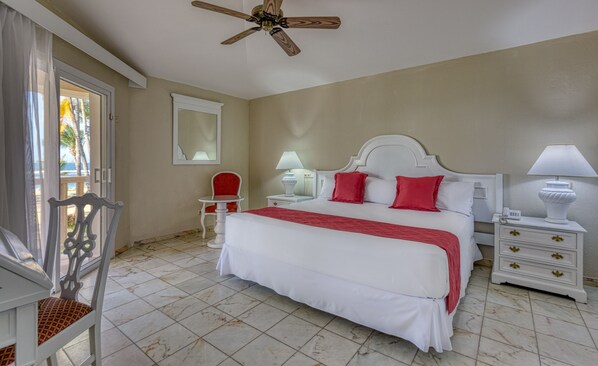 A hotel room with a bed, a chair, a table, a nightstand, a lamp, a ceiling fan, and a window with a view of palm trees.