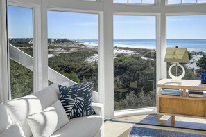 Una habitación con un sofá blanco, una mesita de madera con libros y vistas a la playa y al océano a través de amplios ventanales.