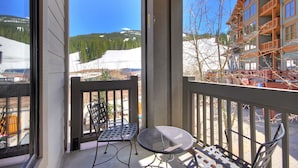 A balcony with two metal chairs and a small round table, overlooking a snowy mountain and a building with multiple balconies.